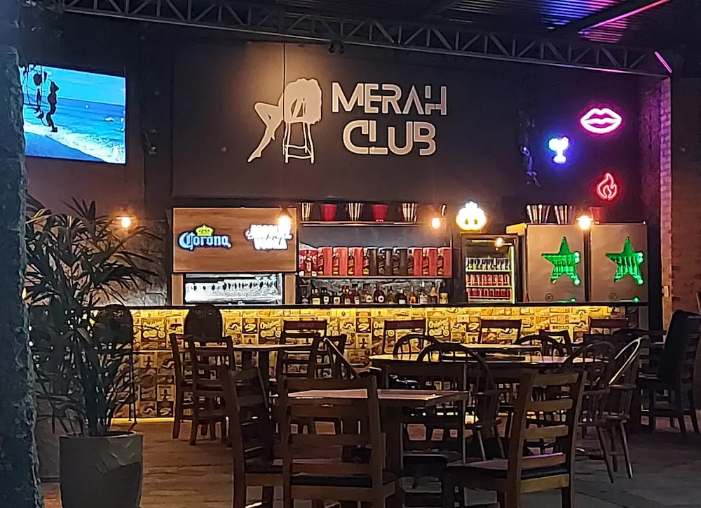 Salão do Merah Club com mesas de madeira e letreiro em neon
